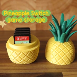 Könnte beinhalten: Ein gelber, ananasförmiger Aufbewahrungsbehälter für Spiele mit einem grünen Blätterdach. Die Basis enthält schwarze Nintendo Switch-Spielkassetten. Der Text "Pineapple Switch game storage" steht darüber, ergänzt durch drei kleine gelbe Sterne.