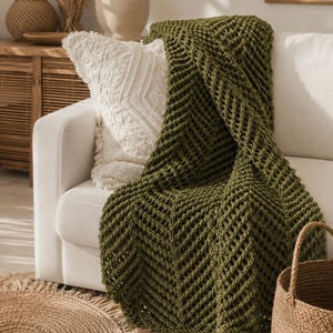Peut inclure: Un plaid en tricot épais de couleur vert olive profond présentant un motif texturé en chevron ou en zigzag. Le plaid est drapé sur l'accoudoir et l'assise d'un canapé blanc, recouvrant partiellement un coussin blanc texturé.