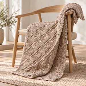 Peut inclure: Un plaid tricoté marron clair avec un motif diagonal texturé drapé sur une chaise en bois. La couverture présente une construction en maille épaisse avec des détails de points visibles et une palette de couleurs neutres et douces.