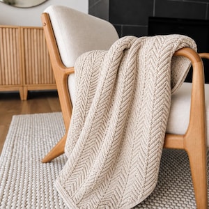 May include: A beige knit throw blanket featuring a prominent herringbone pattern draped over a light-coloured upholstered chair with a wooden frame. The blanket has a textured surface with vertical columns of V-shaped stitches throughout the fabric.