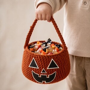 May include: A round, burnt orange crochet basket featuring a jack-o'-lantern face with black felt triangular eyes, a small triangular nose, and a wide, jagged mouth. The basket has a sturdy handle and is filled with assorted wrapped candies.