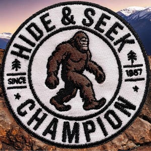 May include: A circular embroidered patch with a white background and black border. It features a brown Bigfoot figure in the center. The text reads HIDE & SEEK CHAMPION, SINCE 1967, with small tree and star icons integrated into the design.