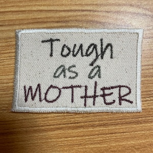 May include: A rectangular patch with the words "Tough as a MOTHER" embroidered in dark green and burgundy thread. The patch has a light beige background and a white border. The patch is likely for clothing or accessories.
