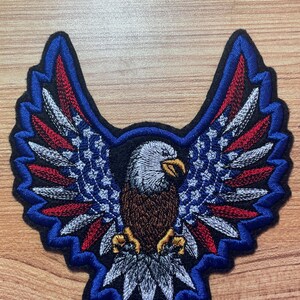 May include: Embroidered patch of a bald eagle with outstretched wings. The eagle's wings are decorated with red, white, and blue stars and stripes, resembling the American flag. The eagle is brown with a white head and yellow talons.