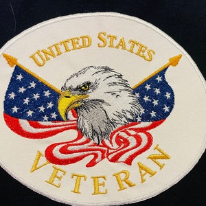 May include: White circular patch with a gold embroidered border. The center features a bald eagle with outstretched wings holding two American flags. The text "UNITED STATES VETERAN" is embroidered in gold around the eagle.