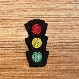 May include: Black embroidered patch of a traffic light with red, yellow, and green circles. The patch is rectangular with a rounded top and bottom. The background is a light brown wood grain.