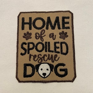 May include: A tan-colored fabric patch with brown stitching and the text "Home of a Spoiled Rescue Dog" in black embroidery. The patch features two paw prints and a white dog face with black eyes and nose.