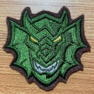 May include: Embroidered patch featuring a green monster face with yellow eyes and a wide, toothy grin. The patch has a brown border and a detailed, textured design, perfect for adding to clothing or accessories.