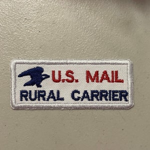 May include: White rectangular patch with blue and red embroidery. The patch features a stylized blue eagle with a red, white, and blue shield, the text "U.S. MAIL" in red, and the text "RURAL CARRIER" in blue.