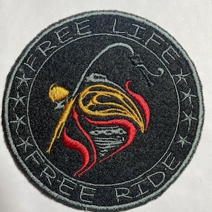 May include: A black, round patch with the words "Free Life Free Ride" embroidered in white around the edge. A stylized image of a flame with red and gold accents is in the center.