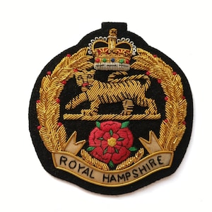 May include: A black fabric badge featuring gold bullion embroidery of a tiger standing above a red rose with green leaves. Above the tiger is a gold crown with red, blue, and white accents. A banner at the bottom reads ROYAL HAMPSHIRE.