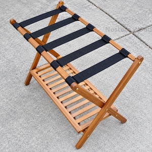 May include: A folding luggage rack constructed from light brown wood with four sturdy black fabric straps stretched across the top frame. The unit features a lower slatted shelf for additional storage and is held together by metal hardware.