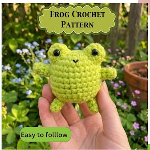 May include: A small, light green crocheted frog toy with black plastic eyes and a stitched mouth. The image includes text that reads FROG CROCHET PATTERN and Easy to follow. The frog has a round body, two small arms, and two small legs.