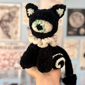 Könnte beinhalten: Ein gehäkeltes Plüschtier in Form einer schwarzen Katze mit einem einzelnen großen zentralen Auge, das eine weiße Iris und mintgrüne Akzente aufweist. Die Katze hat einen weißen Rüschenkragen, weiße Ohrdetails und ein weißes Spiralmuster an der Seite.