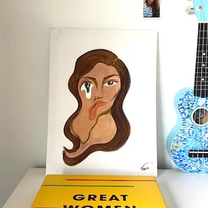 May include: A rectangular canvas painting featuring a stylized portrait of a person with long brown hair, a green eye, and a melting face effect. The painting is signed in the bottom right corner and sits behind a yellow book titled GREAT WOMEN ARTISTS.