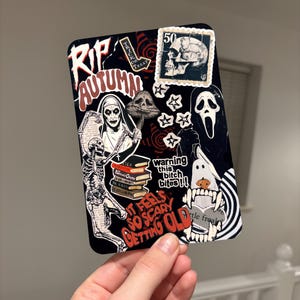 May include: A rectangular sticker sheet featuring a black background with various horror-themed illustrations including a skull, a ghost, a skeleton, a nun, a mushroom, and books. Text includes RIP AUTUMN, TRICK OR TREAT, WARNING THIS BITCH BITES, and IT FEELS SO SCARY GETTING OLD.