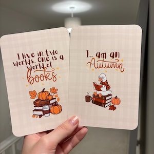 May include: Two rectangular cards with a light beige plaid background. The left card features the text I live in two worlds. one is a world of books above a stack of books, pumpkins, and a coffee cup. The right card shows a goose reading a book.