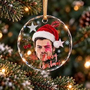 May include: A circular clear glass ornament featuring a portrait of a person wearing a red Santa hat with a pink star graphic on their face. The design includes red cherries, silver and pink stars, snowflakes, a red lip print, and the text Merry Christmas.