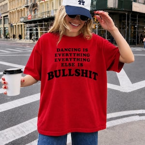 May include: A bright red short-sleeved t-shirt featuring black text that reads DANCING IS EVERYTHING EVERYTHING ELSE IS BULLSHIT. The shirt has a crew neckline and a loose fit, paired with a white and blue baseball cap and a white disposable cup.