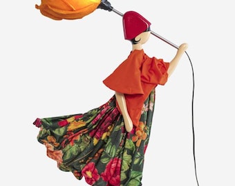 Pode incluir: Um candeeiro decorativo em forma de figura que usa um chapéu vermelho, um top laranja e uma saia verde-escura com padrão floral. A figura segura um grande abajur laranja em forma de flor numa haste metálica sobre uma base.