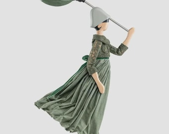 Könnte beinhalten: Eine dekorative Figur, die ein Kleid mit grünem Spitzenoberteil und plissiertem Rock, eine hellgraue Haube und passende Stiefel trägt. Die Figur hält einen grünen Regenschirm mit einem silbernen Metallgriff und steht auf einem Holzsockel.