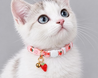 May include: A light pink cat collar featuring a repeating strawberry pattern, a white plastic cat-shaped buckle, and a gold-toned metal ring holding a small gold bell and a red strawberry-shaped charm worn by a white kitten.