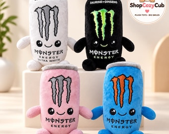 May include: Four plush toys shaped like energy drink cans in white, black, pink, and blue. Each features a smiling face, small arms and legs, and the Monster Energy logo. The white can says ZERO SUGAR, the black can says TAURINE+GINSENG.