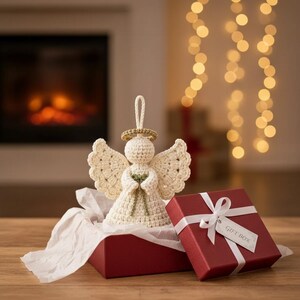 May include: A cream-colored crocheted angel ornament with a gold halo and green ribbon detail, positioned inside an open red gift box with white tissue paper and a white ribbon, featuring a tag with the text GIFT BOX.