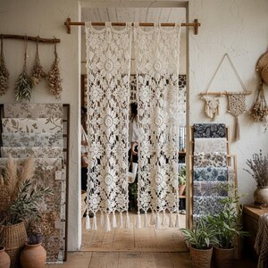 May include: A pair of off-white crochet curtains featuring an intricate floral pattern and a bottom edge finished with tassels. The curtains are suspended from a wooden rod mounted within a doorway, displaying a detailed, open-work textile design.