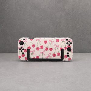Puede incluir: Una consola de juegos portátil con una funda protectora de color crema claro decorada con un patrón repetitivo de cerezas de color rojo brillante y pequeños lazos rosas. El dispositivo incluye botones de control, joysticks y soporte negros.