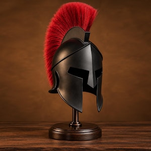 Può includere: Un elmo in stile spartano nero opaco caratterizzato da una cresta rossa alta e prominente fatta di setole rigide. L'elmo è montato su un supporto espositivo circolare in legno scuro, mostrando un design classico con un'apertura facciale a T.