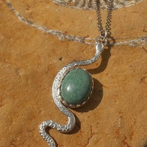 May include: A silver-toned metal pendant shaped like a winding snake with a textured surface, featuring an oval-shaped green stone set in the centre. The pendant hangs from a thin silver-toned metal chain, creating a cohesive jewellery piece.