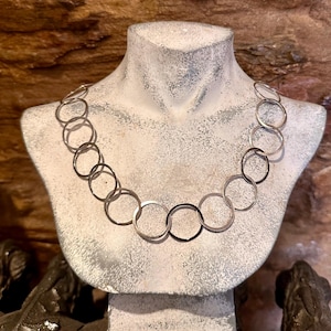 May include: A silver-toned necklace composed of a series of interconnected circular metal rings. The chain features a repeating pattern of linked loops that form a continuous, flexible strand draped across the neck area of a display bust.