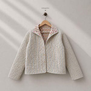 May include: A quilted jacket featuring a cream and dark blue vertical pinstripe pattern on the exterior. The interior lining and the wide collar display a floral print with pink, blue, and green flowers on a tan background. Three buttons close the front.