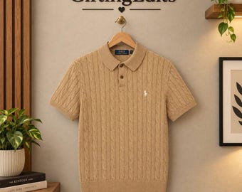 May include: A tan short-sleeve polo shirt featuring a cable knit texture, a collared neckline with two buttons, and a small white embroidered logo on the left chest. The shirt is displayed on a wooden hanger below the text GiftingEdits.