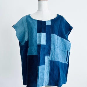 May include: A short-sleeved top featuring a patchwork design composed of various rectangular fabric panels in multiple shades of blue, ranging from light sky blue to deep navy. The garment has a wide, rounded neckline and a loose, boxy fit.