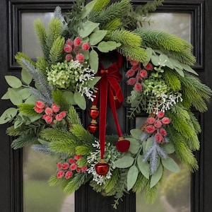 May include: A circular wreath featuring green pine needles, eucalyptus leaves, and clusters of red berries. The arrangement is accented with white floral sprigs and a deep red velvet ribbon tied in a bow, with three small red bell ornaments hanging down.