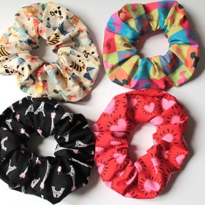 May include: A set of four fabric hair scrunchies. One is cream with animal prints, one features a colourful grid with dog silhouettes, one is black with white Eiffel Tower patterns, and one is red with pink heart and sunburst designs.