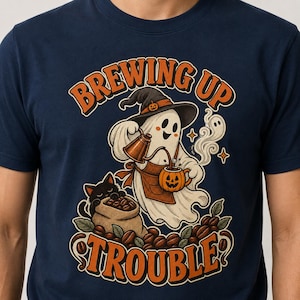 May include: A navy blue t-shirt featuring a graphic of a ghost wearing a witch hat and apron, pouring liquid from a copper kettle into a pumpkin mug. A black cat sits by a burlap sack of coffee beans. The text reads BREWING UP TROUBLE.