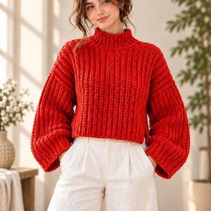 Puede incluir: Un suéter de punto grueso en color rojo vibrante con cuello alto, hombros caídos y mangas anchas y voluminosas. La prenda presenta una textura acanalada vertical en toda su superficie, un dobladillo corto con aberturas laterales y se combina con pantalones blancos.