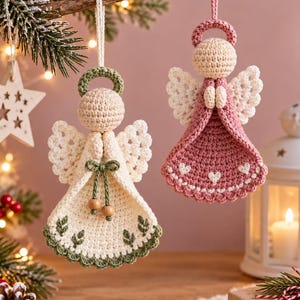 May include: Two handmade crochet angel ornaments hanging from a branch. One angel is cream-colored with green accents and wooden beads, while the other is dusty pink with white heart details. Both feature circular halos and delicate wings.