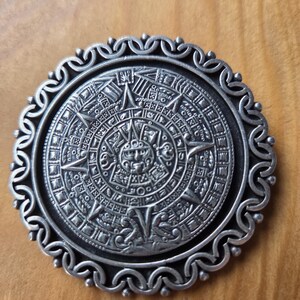 May include: A circular silver-toned metal medallion featuring a detailed relief of the Aztec sun stone calendar. The central face is surrounded by concentric rings of intricate glyphs and symbols, enclosed by a decorative scalloped border.