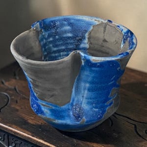 Puede incluir: Un cuenco de cerámica hecho a mano con un borde irregular y ondulado, decorado con un esmalte azul intenso que gotea por los lados. Las superficies interior y exterior presentan un acabado de arcilla gris mate con textura visible.