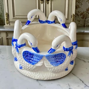 Peut inclure: Un bol en céramique couleur crème doté d'une base circulaire ornée de plusieurs figurines d'oies sculptées sur le pourtour. Chaque oie présente un bec peint en bleu, des détails d'ailes bleus et un ruban bleu noué au cou.