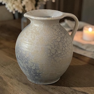 May include: A round ceramic pitcher featuring a wide mouth, a curved handle, and a mottled glaze finish. The surface displays a blend of cream, beige, and soft grey tones with a textured, speckled pattern that covers the entire exterior body.