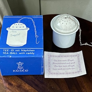 Może przedstawiać: Biały ceramiczny zaparzacz do herbaty z podstawką, zaprezentowany w niebieskim pudełku z napisem "TEE-EI mit Näpfchen TEA-BALL with caddy" oraz "KAISER". Dołączona jest mała karteczka z niemieckim tekstem "Du bist mein Engel. Beschützend und groß. Du bist mein Engel. lass mich nie mehr los..."