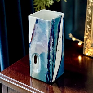 Può includere: Un vaso rettangolare in ceramica con base bianca e gocce di vernice astratta in tonalità blu e viola in stile acquerello. Il vaso presenta una finitura liscia, un'apertura ovale su un lato e un pannello bianco decorato con piccoli punti neri.