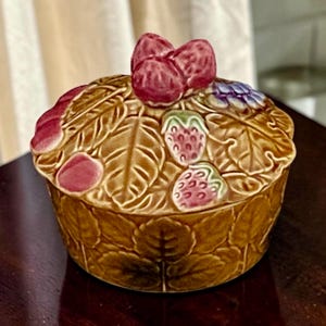 May include: A round ceramic lidded jar featuring a brown leaf-textured surface. The lid is adorned with sculpted red strawberries and green leaves, with a cluster of three strawberries forming the handle. The base also displays embossed leaves.