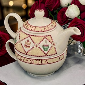 Può includere: Un set da tè individuale in ceramica color crema composto da una teiera impilata su una tazza. Il set è decorato con un motivo a rombi contenente illustrazioni di focaccine e marmellata, con la scritta CREAM TEA in lettere rosso scuro.