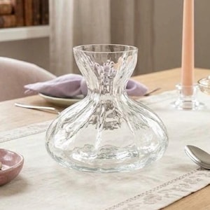 May include: A clear glass vase with a wide, rounded base and a narrow, fluted neck. The glass features a textured, rippled surface design that catches the light, creating a series of vertical indentations around the body of the vessel.